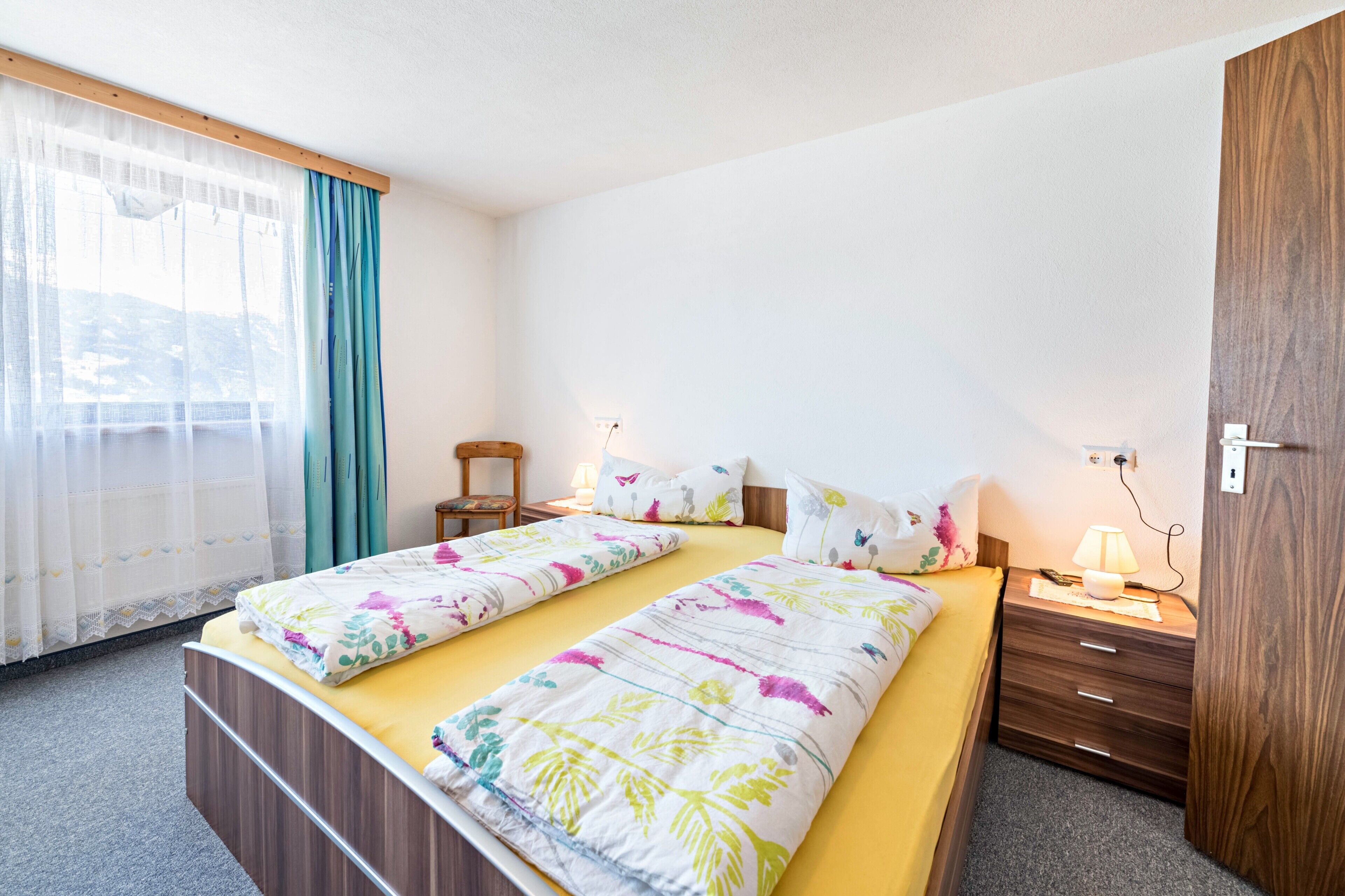 A double bed with floral bedding, a bedside table with a lamp, a wooden nightstand, and a chair.