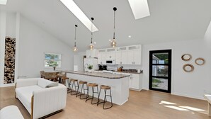 Una cocina moderna con armarios blancos, una isla central con taburetes, iluminación colgante y un ventanal amplio.
