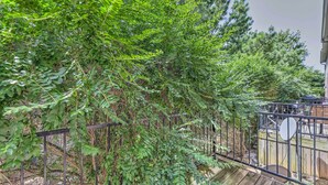 A wooden deck with a metal railing, surrounded by dense green foliage. - Charming Springdale Townhome ~ 5 Mi to Dtwn! (Springdale)