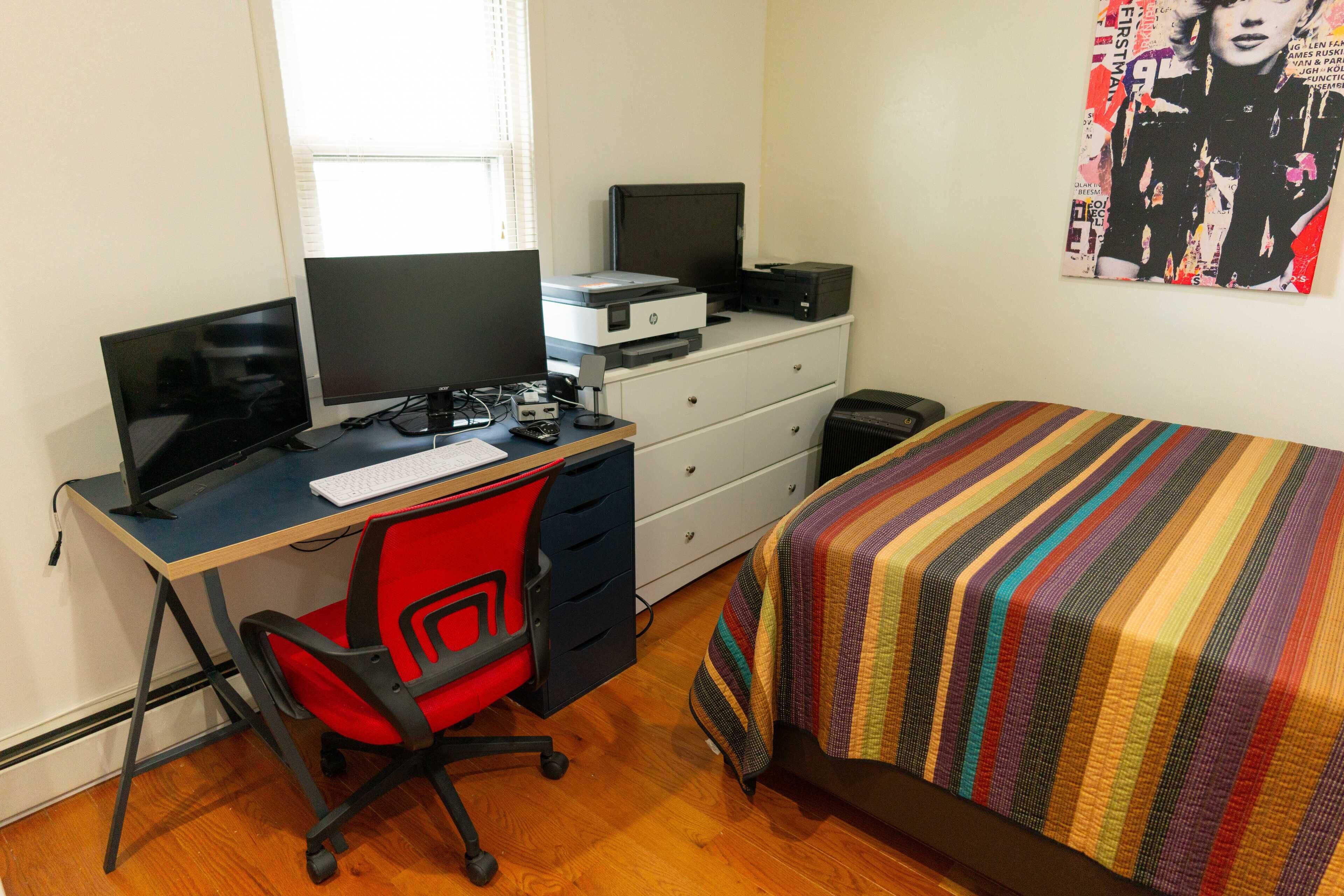 Une chambre à coucher avec un lit, un bureau avec un ordinateur, une chaise, une commode et une affiche accrochée au mur.
