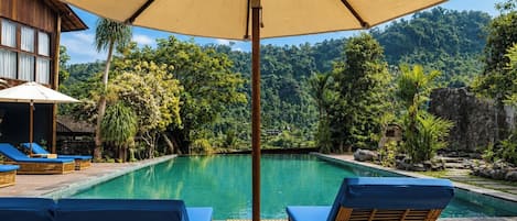 A pool area with lounge chairs and umbrellas, surrounded by lush greenery and a mountainous backdrop.