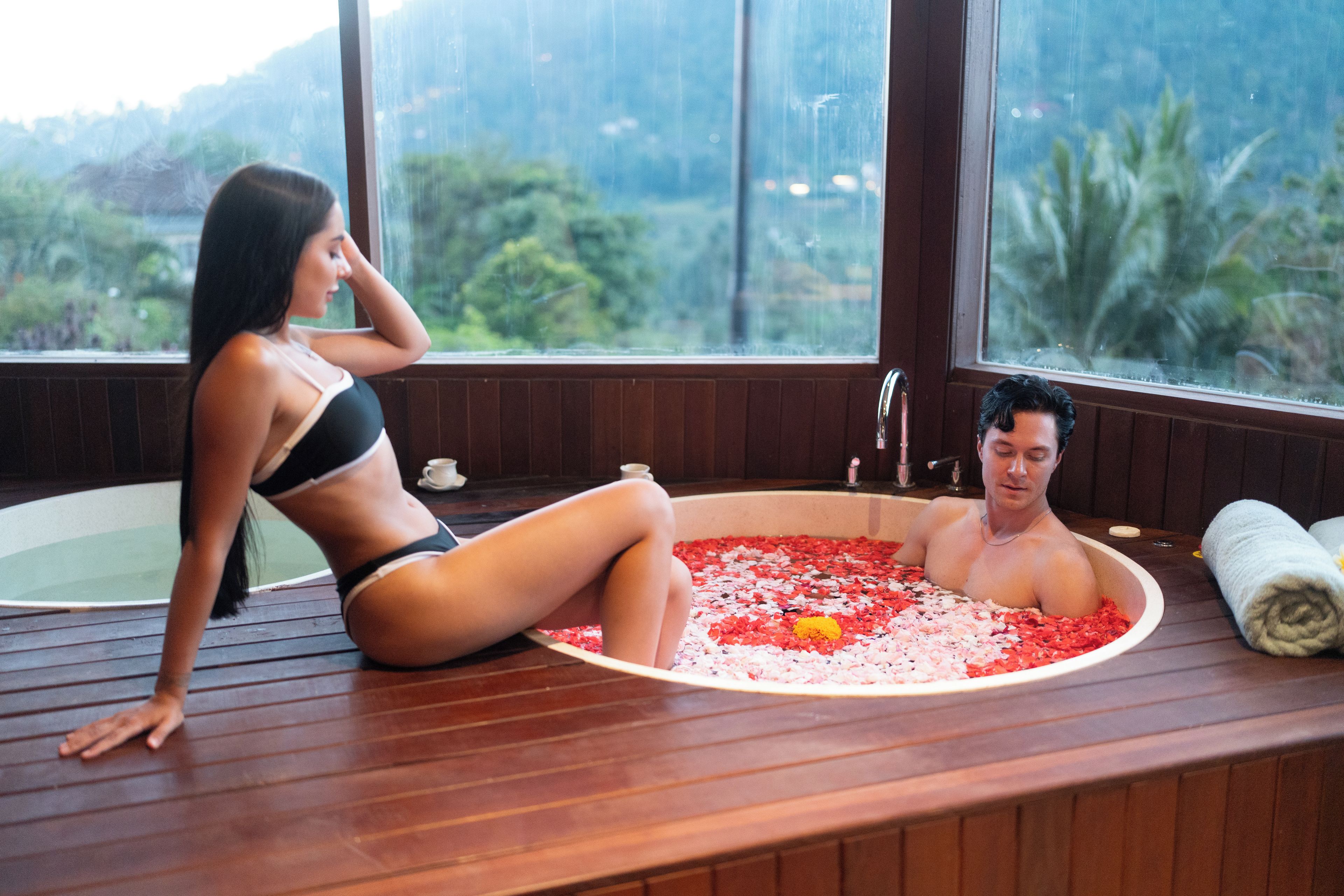 Un hombre y una mujer en una habitación de madera con un ventanal grande, sentados en una bañera llena de agua y pétalos de flores.