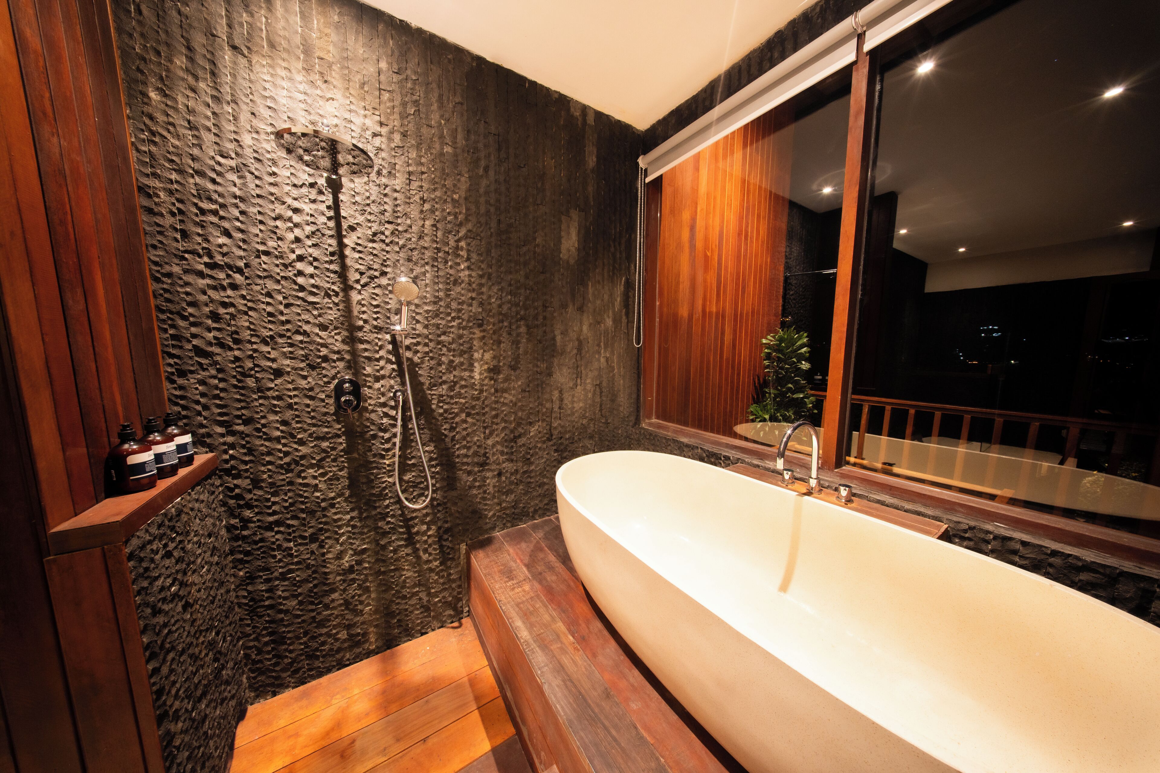 Un baño moderno con pared de piedra, estante de madera con botellas, una bañera grande y ventana con vistas.
