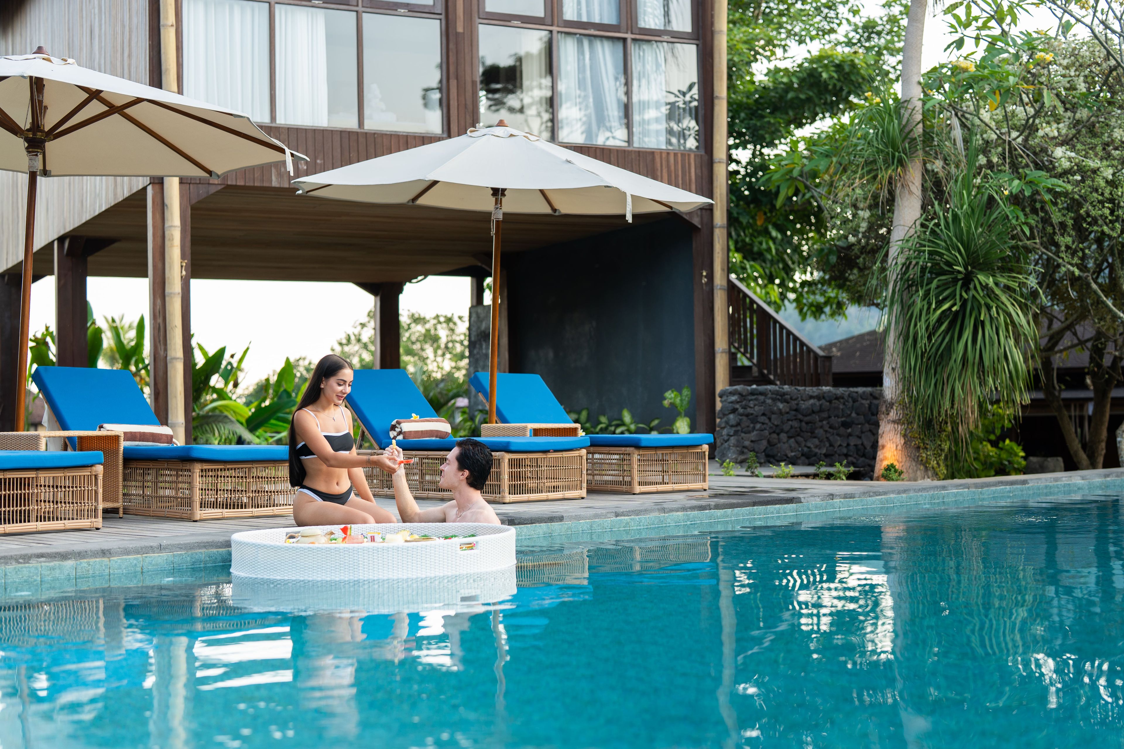 Zona junto a la piscina con sillones de descanso y sombrillas, una piscina y un edificio con amplias ventanas al fondo.
