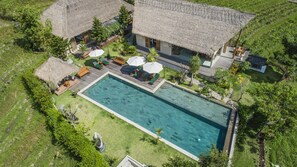 Vue aérienne d’un complexe hôtelier doté d’une grande piscine, de bâtiments à toits de chaume et d’une végétation luxuriante.