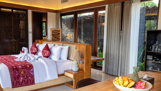 Habitación de hotel con cama, mesita de noche, lámpara y un cuenco con fruta sobre una mesa de madera.