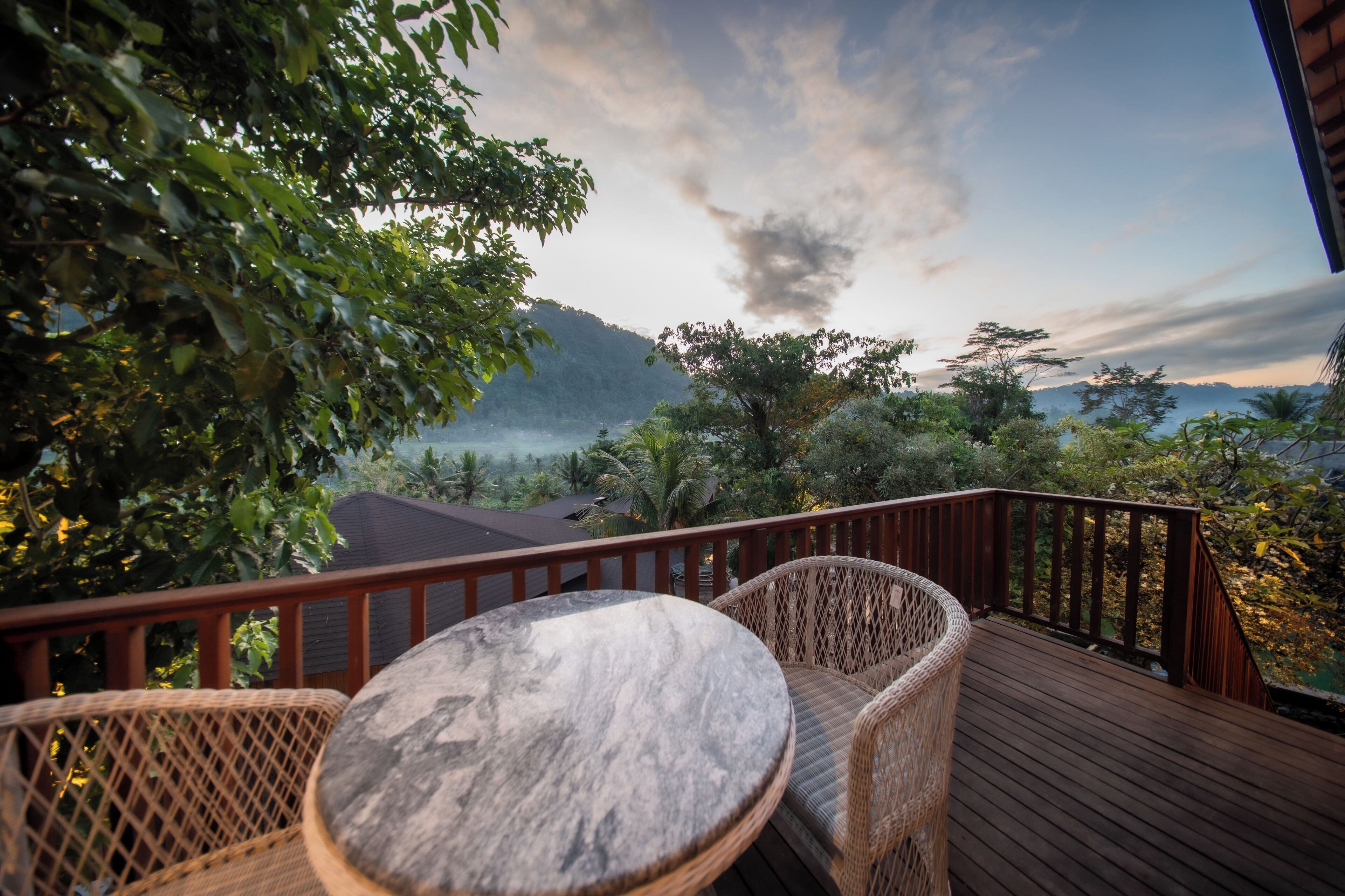Una terraza de madera con mobiliario de mimbre y una mesa de mármol, con vistas a un paisaje montañoso.