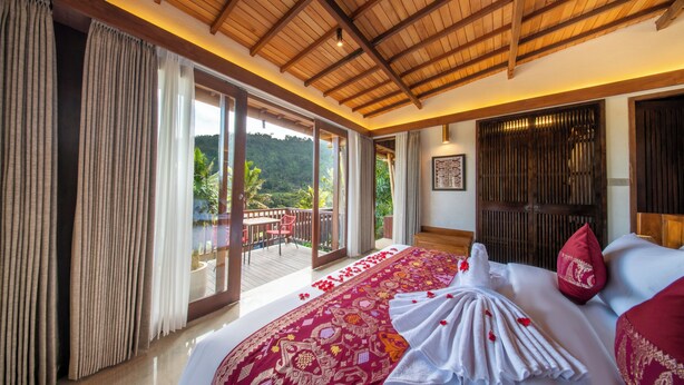 A bedroom with a large bed, a balcony, and a view of a lush landscape.