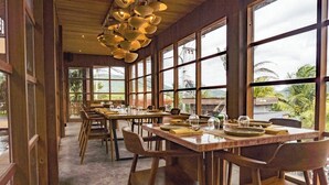 A restaurant with wooden tables and chairs, large windows, and a view of greenery.