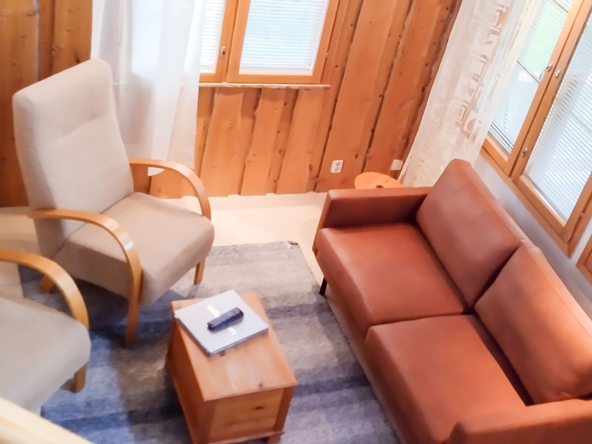 Et værelse med trægulv, en beige sofa, en træstol, et lille træbord med en fjernbetjening og et vindue med rullegardiner.