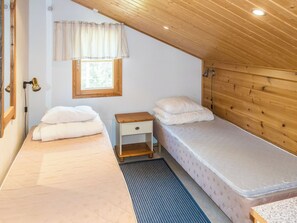 Une petite pièce accueillante avec un plafond en pente, une fenêtre ornée d’un rideau léger, une table de chevet en bois, deux lits avec du linge de lit blanc et un petit tapis.