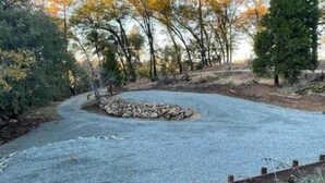 A gravel parking area with a circular stone feature, surrounded by trees and a wooden fence.
