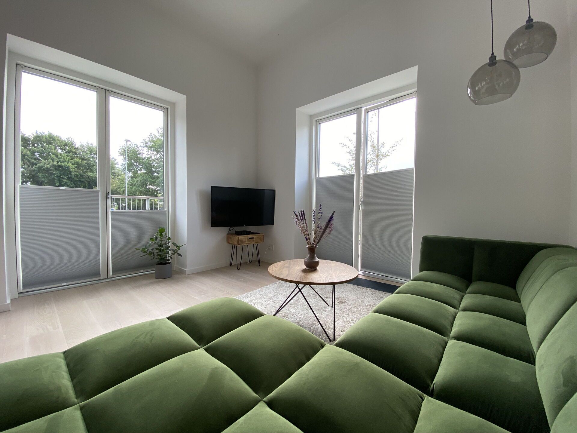 A modern living room with a green sectional sofa, a flat-screen TV, a small wooden table with a vase, and large windows.