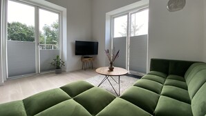 Una sala de estar moderna con un sofá seccional verde, un televisor de pantalla plana, una pequeña mesa de madera con un florero y ventanas amplias.