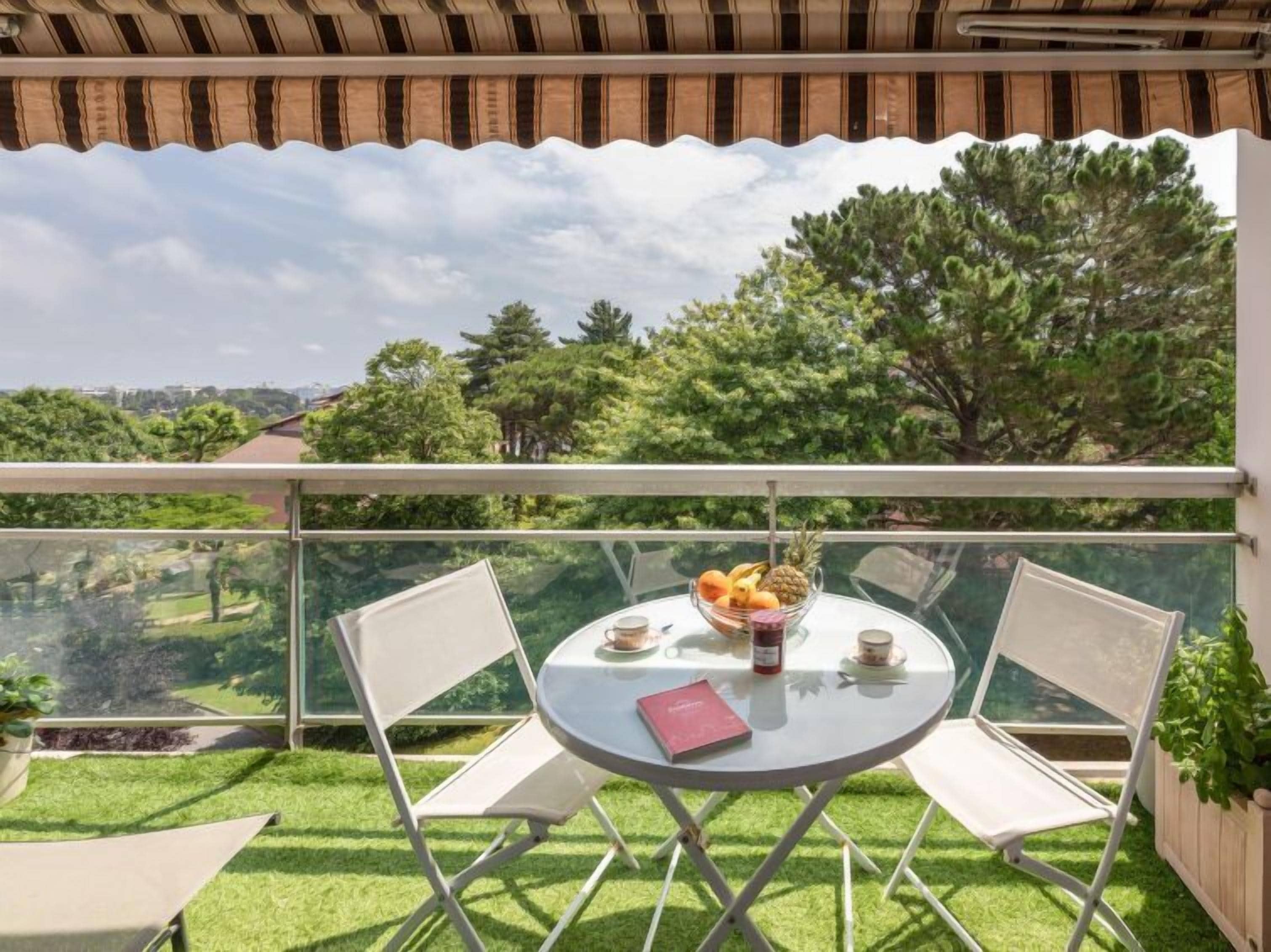 A balcony with a glass table, two chairs, a bowl of fruit, and a book.