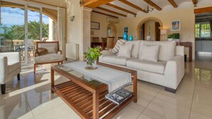 A spacious living room with a large sofa, a coffee table, and a glass-top table. There is a wooden ceiling with exposed beams, and a view of the outdoors through large windows. - Isabelle by Interhome (Jávea)