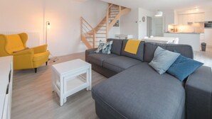 Uma sala de estar moderna com um sofá cinza, uma poltrona amarela e uma mesa de centro branca. Uma escada leva a um andar superior.