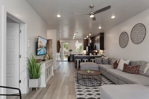 A modern living room with a sofa, coffee table, and a TV. There is a kitchen area with a bar and stools. - Red Desert Loft Resort 37| Pickleball Courts, Foosball, Resort Pool with Waterslide & Jacuzzi! (Saint George)