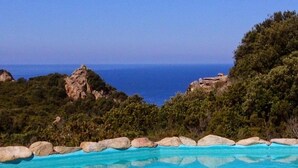 Piscina azul límpida com borda rochosa, rodeada por vegetação exuberante e com vista para a paisagem costeira.