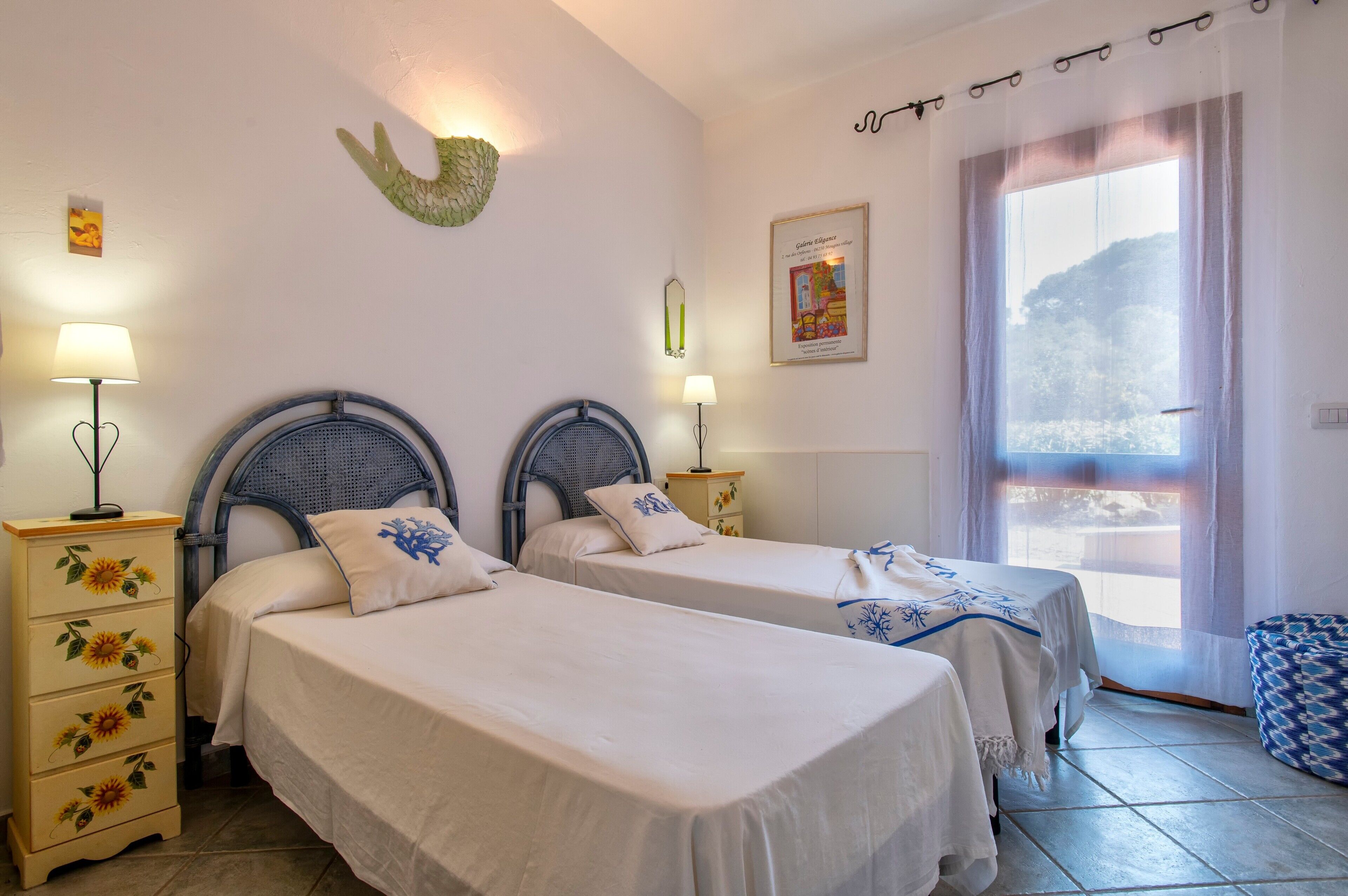 Una habitación con dos camas, una mesita de noche, una lámpara, un adorno en la pared, un cuadro enmarco y una ventana con vistas.