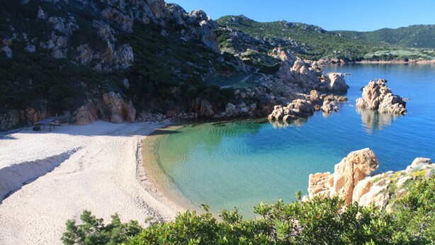 Uma praia com águas cristalinas, afloramentos rochosos e vegetação.