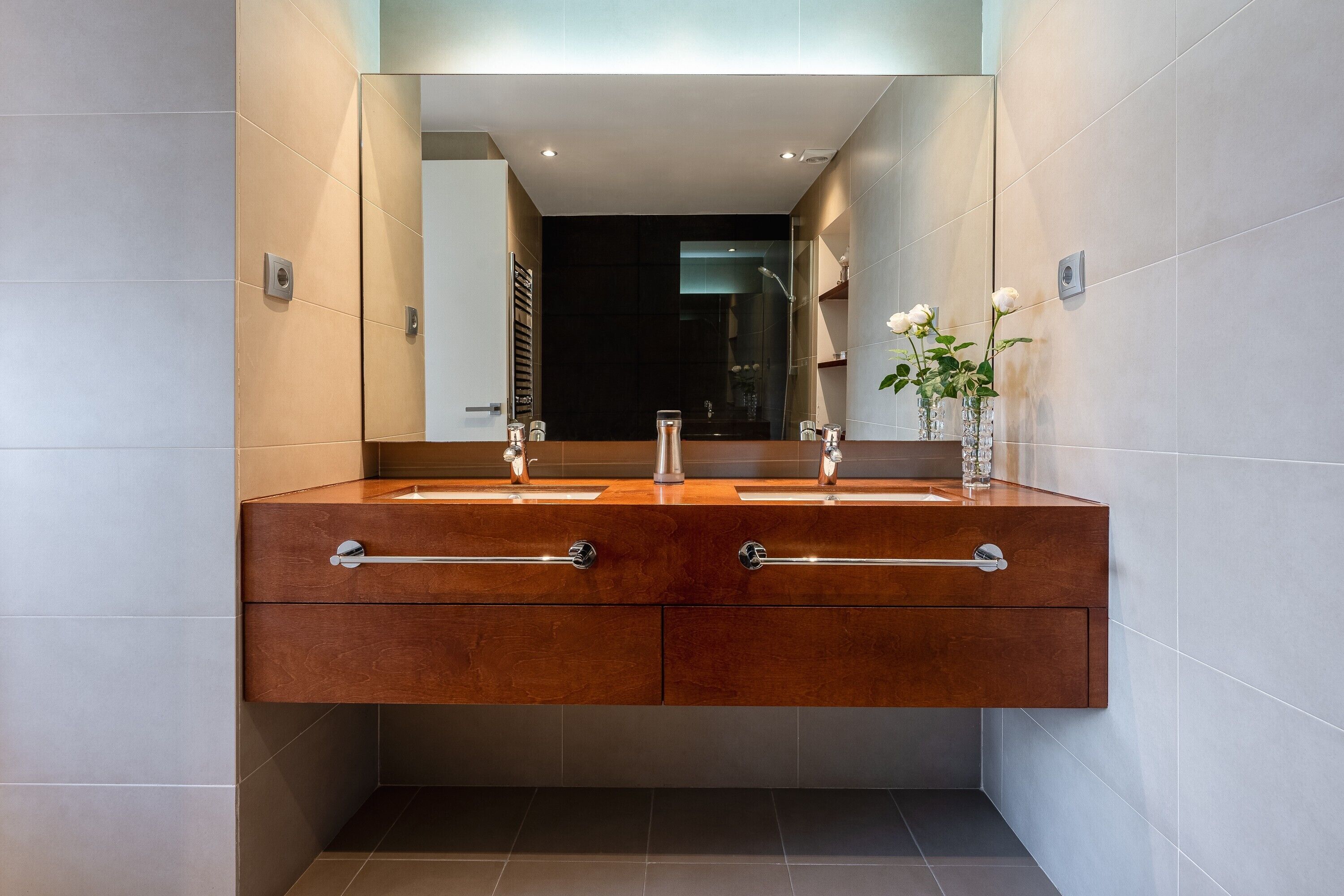 A modern bathroom with a double vanity, a large mirror, and a vase with flowers.