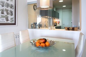 A dining table with a glass top and a metal bowl containing fruit. There are white chairs around the table.