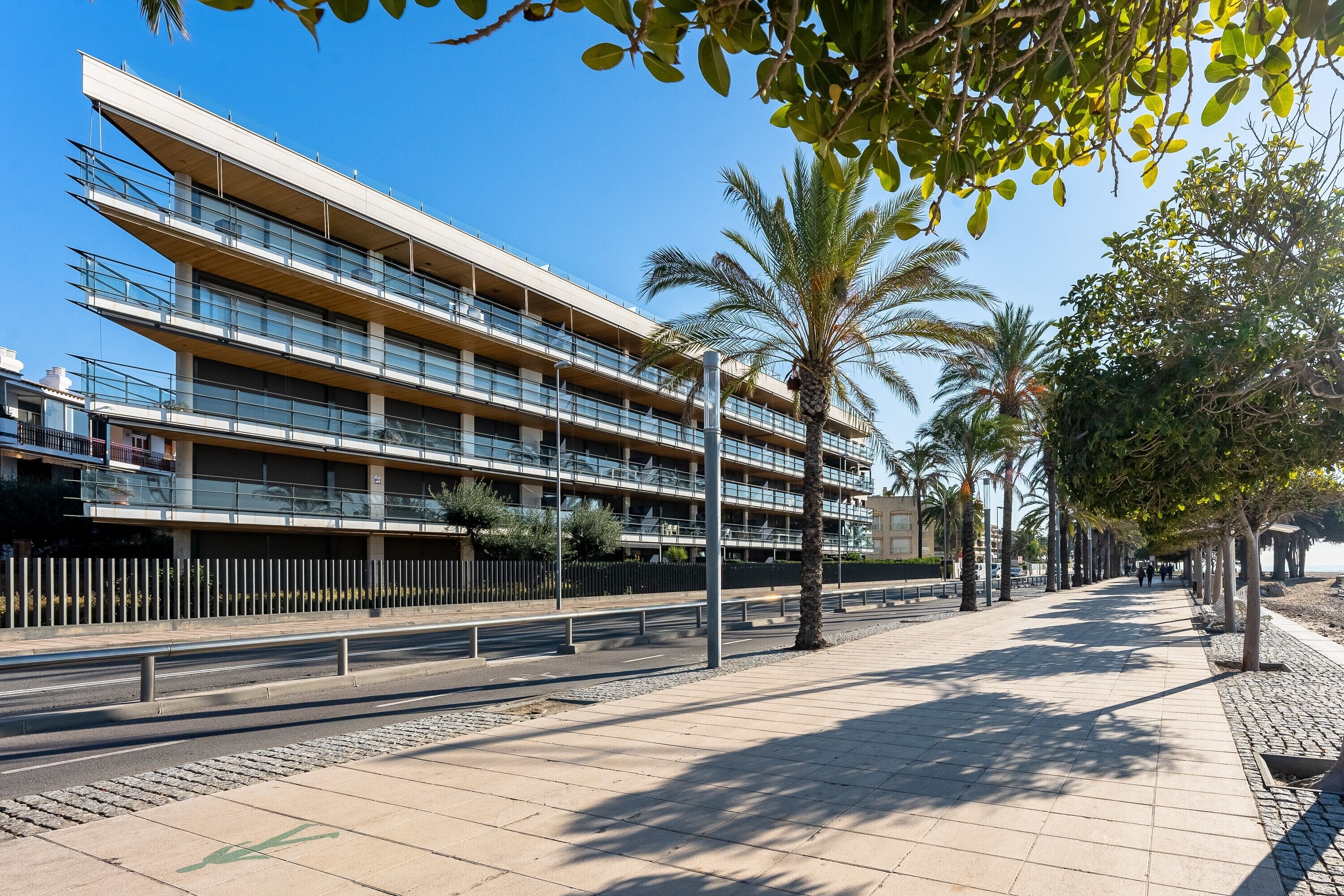 A multi-story building with balconies, a paved walkway, and palm trees.