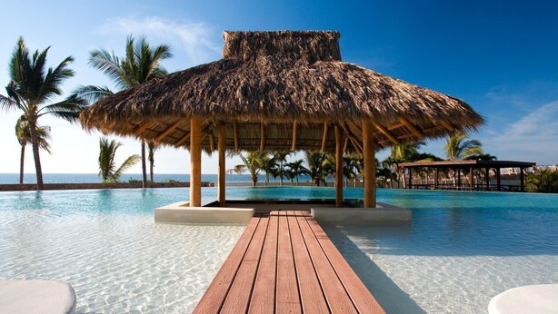 A pool area with a wooden deck leading to a thatched-roof structure, surrounded by palm trees and a clear view of the ocean.