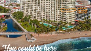 A high-rise hotel with multiple balconies, a beach, and a river.