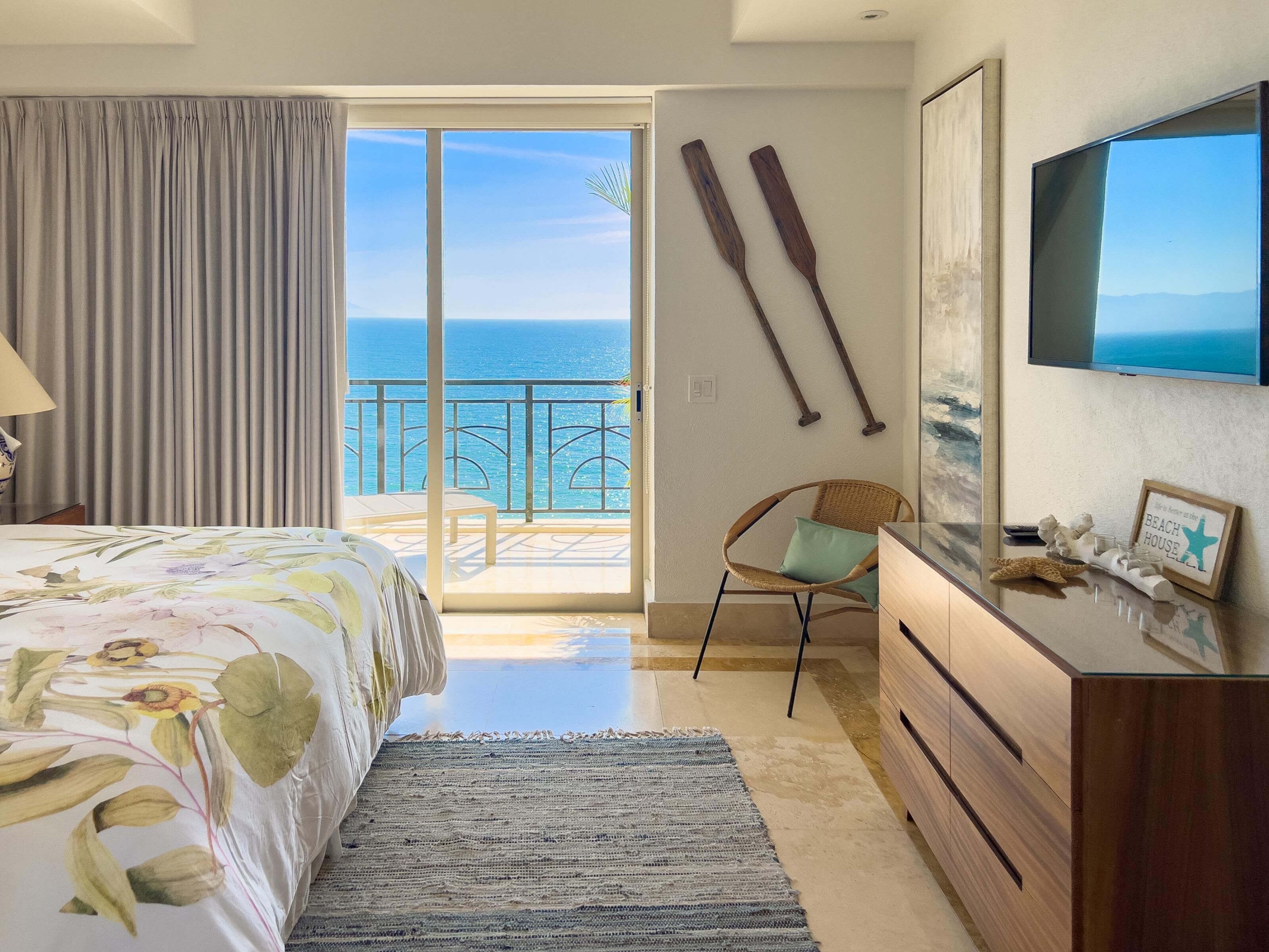A bedroom with a bed, a dresser, a chair, a TV, and a view of the ocean.