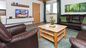 Olohuoneessa on ruskeat nahkasohvat, puinen sohvapöytä, litteänäyttöinen televisio ja suuri ikkuna, jossa on verhot.