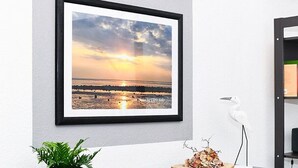 Een ingelijste foto van een zonsondergang boven een strand, een potplant en een decoratieve vogel op een tafel.
