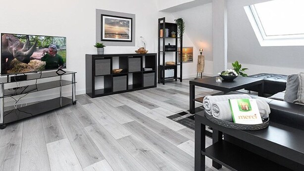 Een moderne woonkamer met een flatscreen televisie, een zwarte tv-meubel, een eettafel met een mand, en een raam met natuurlijk licht.