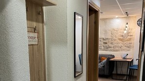 En gang med en træbænk, et indrammet billede og et skilt med teksten 'Welcome'. I baggrunden ses et spiseområde med et bord og stole.