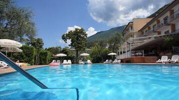 A swimming pool with lounge chairs and umbrellas, surrounded by trees and a building with balconies.