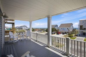 A covered porch with white railings, outdoor seating, and a view of the ocean and neighboring houses.