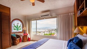 A hotel room with a large bed, a view of a cityscape, a wooden ceiling fan, and a window with curtains.