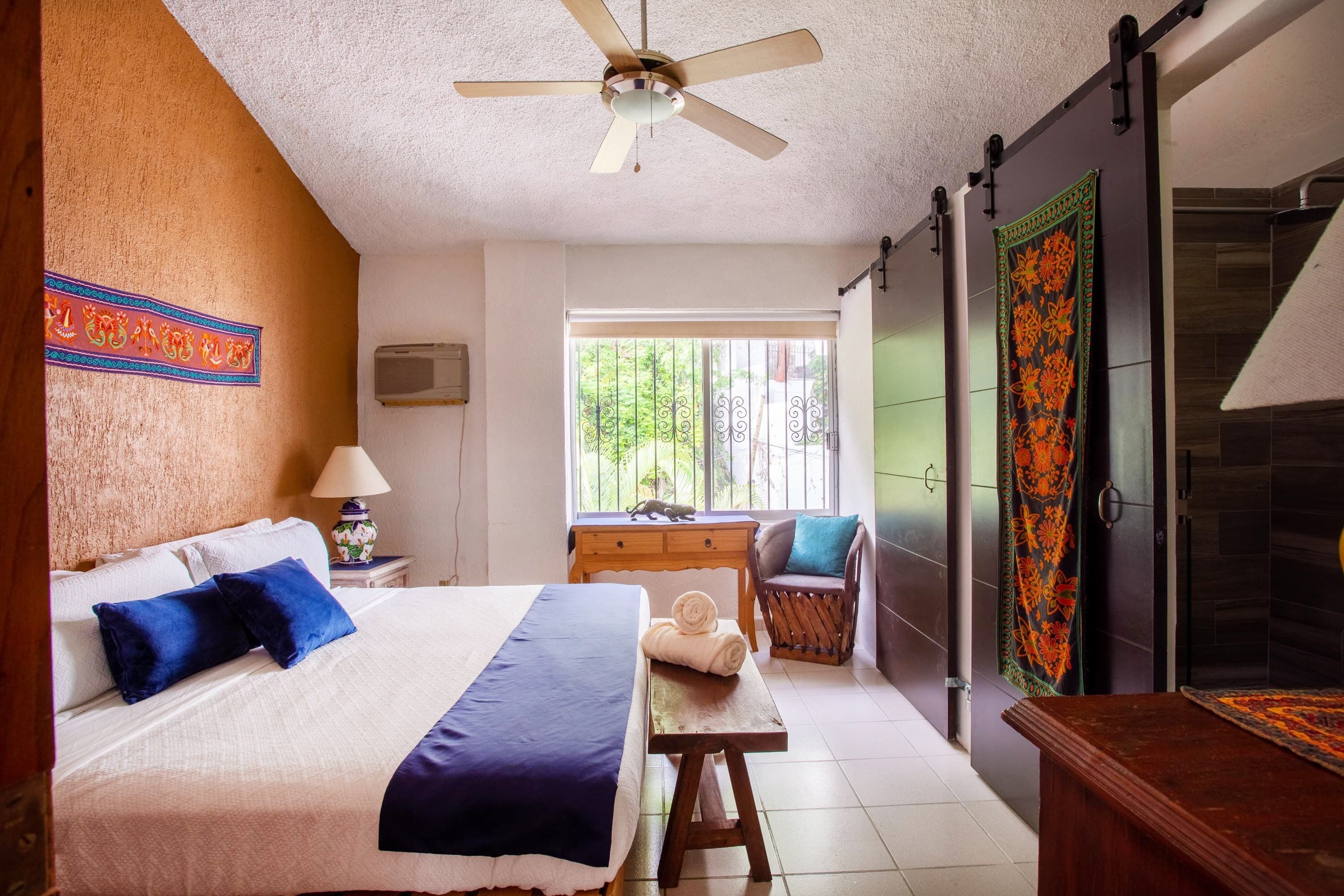 A hotel room with a bed, a desk, a chair, a ceiling fan, a window with a view of greenery, and a decorative tapestry.