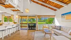 A spacious living area with a wooden ceiling, a dining area with white stools, and a large sliding glass door leading to a balcony with a view of greenery. - Beautiful 2 Br 2.5 Ba Condo Golf Course View, Pool, Hot Tub (209AB) (Princeville)