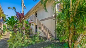 A building with a white exterior, a wooden staircase, and lush tropical vegetation. - Bright and Beautiful Princeville Studio, Private Lanai, Pool (209B) (Princeville)
