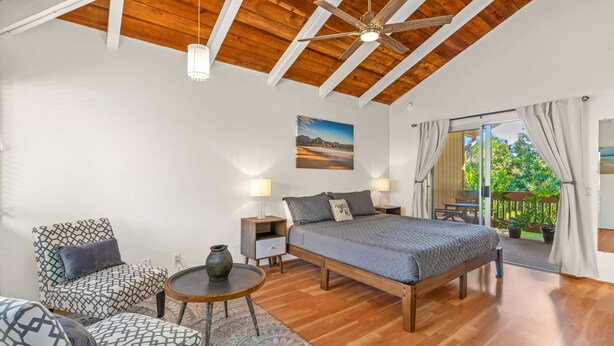 A bedroom with a wooden ceiling, a bed with a gray blanket, two nightstands, a chair, a small table, and a view of the outdoors through a sliding door. - Bright and Beautiful Princeville Studio, Private Lanai, Pool (209B) (Princeville)