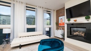 A modern living room with a sofa, a blue armchair, a fireplace, a TV, and large windows with curtains. - Private Balcony | Firepit | Portable Bar (Ketchum)