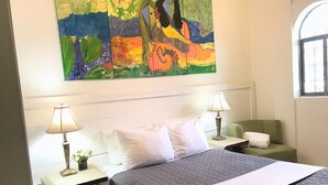 Quarto de hotel com uma cama grande, duas mesas de cabeceira com luminárias, uma poltrona verde e uma pintura abstrata colorida sobre a cama.