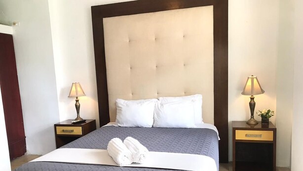 Cama bem arrumada com um edredom cinza, duas almofadas brancas e toalhas enroladas sobre um colchão branco. Há duas mesas de cabeceira com abajures e um pequeno vaso com flores em uma delas.