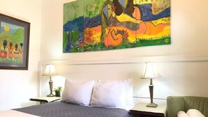 Quarto de hotel com uma cama, duas luminárias de cabeceira, uma poltrona verde e um grande quadro abstrato na parede.