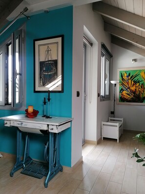 A room with a teal accent wall, a framed black and white maritime painting, a small white table with a candle, and a colorful abstract painting.