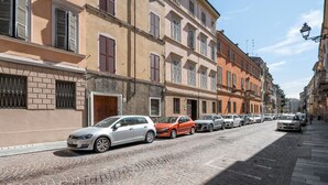 Una strada acciottolata fiancheggiata da edifici storici, auto parcheggiate e un cielo sereno.