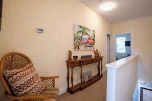 A living room with a wicker chair, a console table with decorative items, a painting of a car and palm trees, and a white door leading to another room. - Mellow Yellow (Carolina Beach)