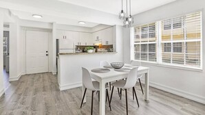 Cozinha moderna com armários brancos, área de jantar com mesa e cadeiras brancas e uma grande janela com persianas.